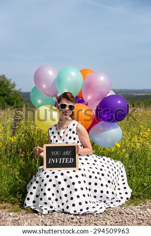 young women in a 50Ã?Â´s dress and colorful balloons an the road, board with text you are invited, concept invitation