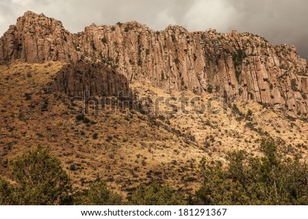 Scenic with imposing wall of column rocks above steep slope/High Cliff Wall of Steep Volcanic Rock Pinnacles Above Steep Mountainside on Gray Cloudy Day/Columns of rock