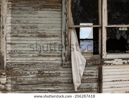 Old white curtain drapes out of open sash window in vintage wooden board house/Worn White Curtain hangs out of Open Old Sash Window/Weathered white curtain hangs through open part of old sash window