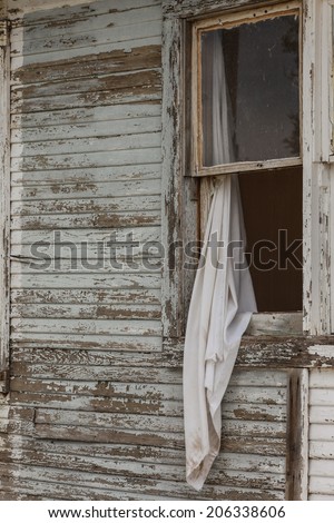 Old curtain drapes from open old sash window in vintage lapboard siding/Still-Life of Old White Curtain out of Derelict Open Sash Window in Clapboard/Old curtain hangs from window in weathered siding