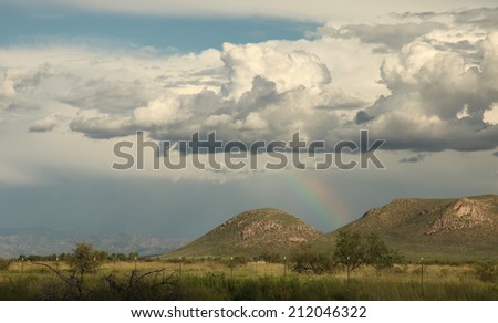 Part of rainbow shows delicate colors beneath storm clouds over landscape with hills and wild vegetation/Rainbow under Clumped Clouds/Part of rainbow with soft colors under cumulus in rural landscape