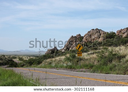 Yellow highway sign showing curving road in rocky scenic/Yellow Roadside Sign with Cautioning Curvy Arrow in Rocky Landscape/Yellow sign on roadside warning of curving road