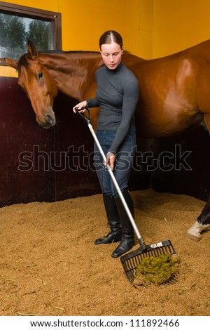 Woman cleans horse box by horse fork, vertical format
