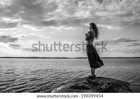 Beautiful young woman looks at the horizon on shore, wind blowing and cloudy sky - black and white image