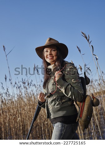 Waterfowl hunting, smiling female hunter carry a shotgun and a decoys, reeds and blue sky on background