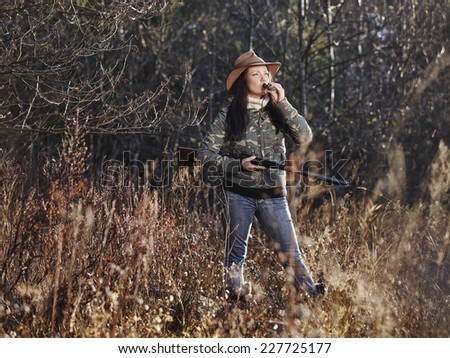 Waterfowl hunting, the female hunter carry a shotgun and she use a duck call, autumnal bushes on background