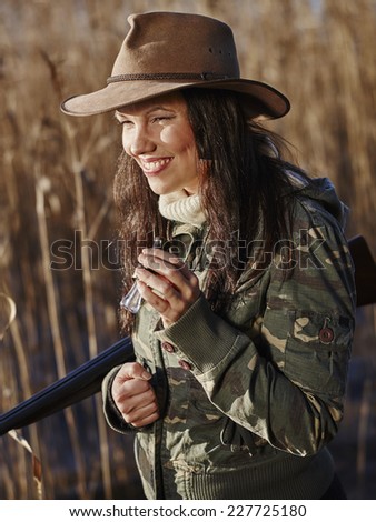 Waterfowl hunting, smiling female hunter carry a shotgun and she use a duck call, shore and reeds on background