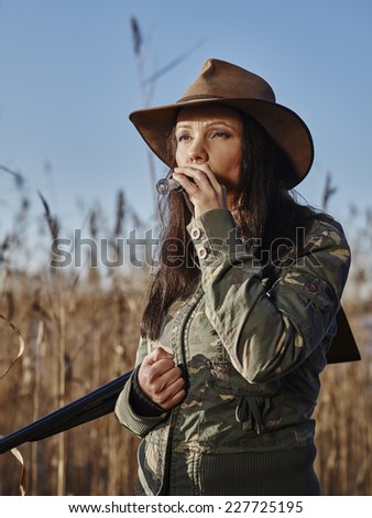 Waterfowl hunting, the female hunter carry a shotgun and she use a duck call, blue sky and reeds on background