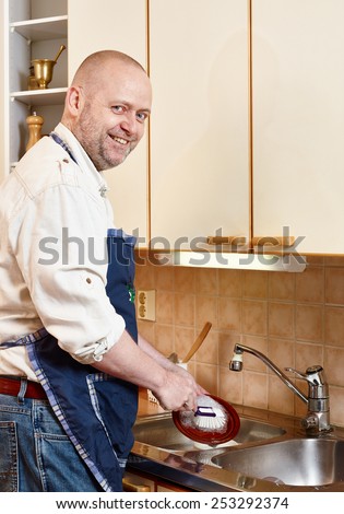 Homeworks, smiling man washes dishes by hand and he looks towards to camera