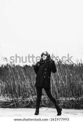 Fashion woman wearing a winter coat and fur cap and she posing front of the reeds, cold rainy weather, full length black and white image
