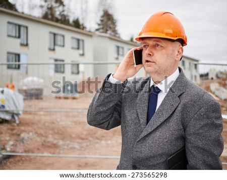 Male engineer uses cell phone, he wearing the suit and the hard hat, house construction site on background