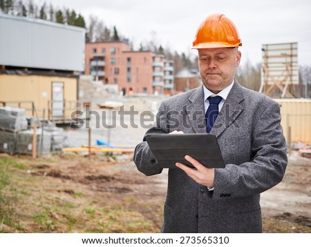 Male engineer uses tablet computer, he wearing the suit and the hard hat, house construction site on background