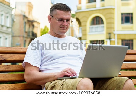 Middle-aged man in glasses sitting on a wooden bench in an urban environment working on his laptop