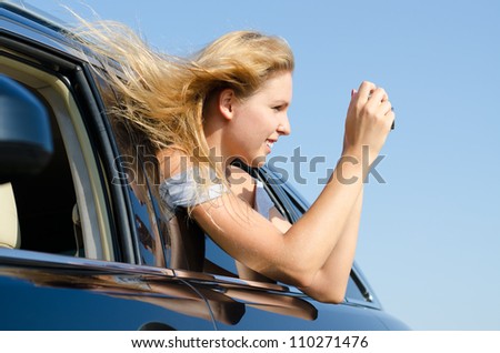 Young woman with her blonde hair blowing in the breeze leaning out of a car taking photographs