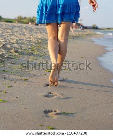 The legs of a barefoot woman walking away from the camera across wet sand on a beach leaving a line of footprints