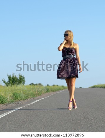 Fashionable woman wearing sunglasses and a party dress walking along a country road in the sunshine