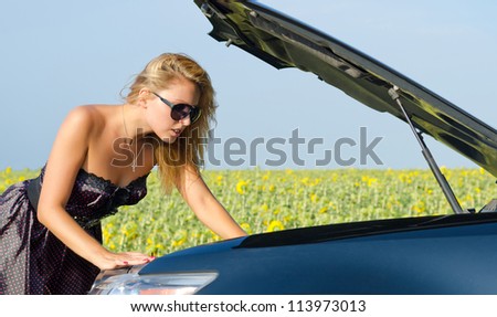 Beautiful woman leaning over with her hand in the engine compartment looking at her car engine near a field of sunflowers