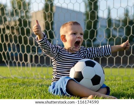 Small boy with a football or soccer ball on his lap sitting in the goalposts shouting with glee and giving a thumbs up gesture