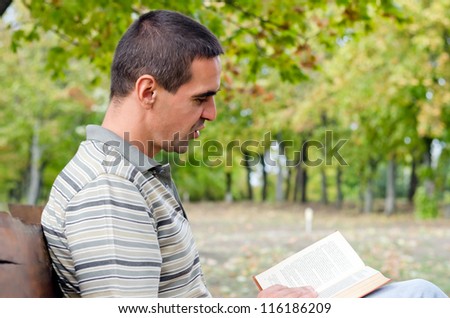Man sitting sideways to the camera on a wooden bench enjoying reading a book outdoors with woodland trees and copyspace
