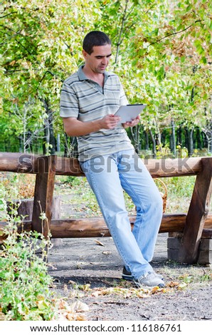 Attractive middle-aged man in jeans leaning on a timber fence in woodland with a tablet in his hands reading the information on the screen
