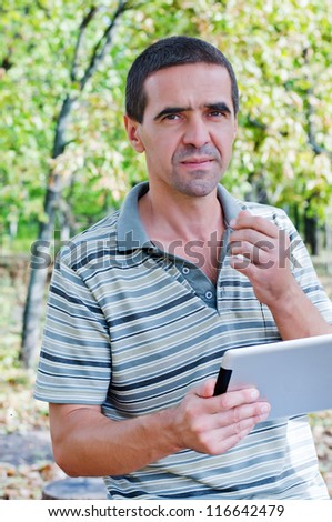 Serious attractive middle-aged man standing outdoors amongst trees using a tablet