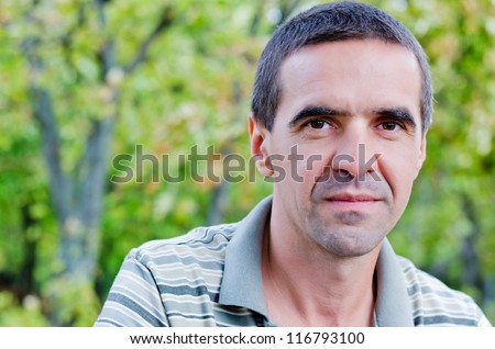 Closeup portrait of an attractive serious middle-aged man looking directly into the camera standing outdoors amongst trees with copyspace