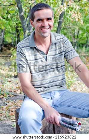 Laughing attractive middle-aged man sitting on a tree stump in the countryside with a rolled up magazine in his hand