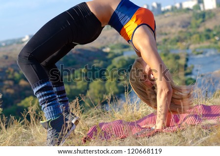Supple athletic woman bending back and arching her back so that both her feet and hands are in contact with ground in open countryside overlooking a river