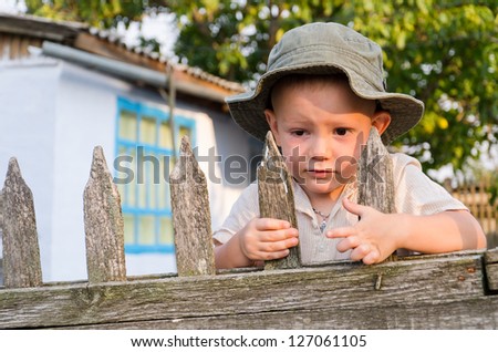 Cute little boy with a worried expression clinging to an old slatted wooden fence in his sunhat waiting for Dad to come home
