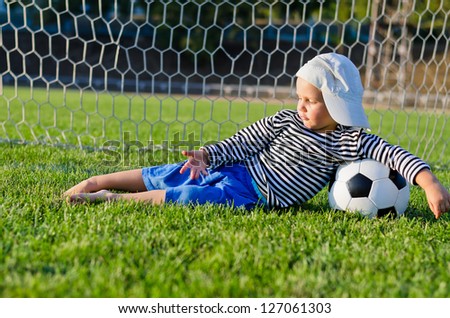 Small barefoot boy in a cap lying relaxing on the green grass in the goalposts with his arm draped over a soccer ball