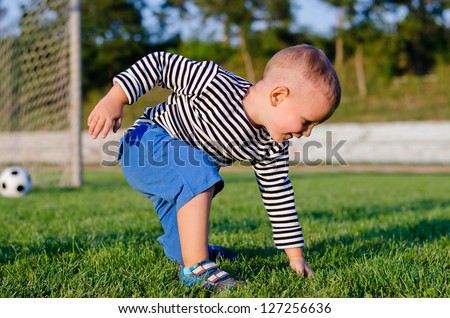 Cute little boy on a soccer field kneeling down to touch the green grass with the goalposts and ball visible behind him