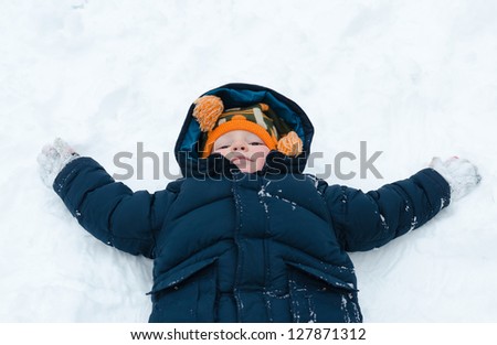 Little boy lying on his back in the snow in his winter jacket playing snow angels with his arms outspread