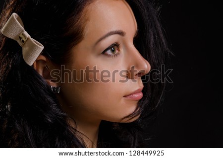 Profile of a beautiful young woman with a bow in her long brunette hair staring into the distance in shadows on a dark studio background