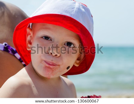Young boy eating watermelon at the seaside looking at the camera with his face full of pink juice and a pip stuck to his cheek