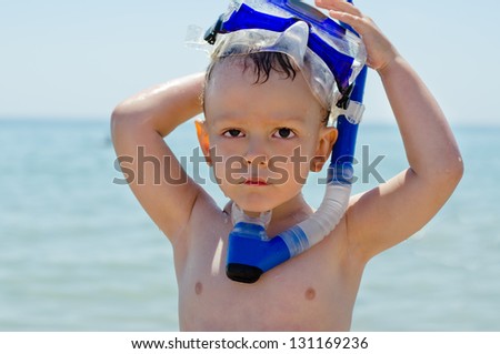 Cute little boy out snorkeling raising his snorkel and goggles to the top of his head as he stands in front of the ocean looking at the camera