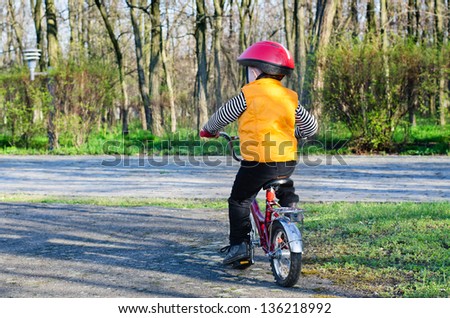 Little boy in a colourful safety helmet and visibility jacket riding his bicycle away from the viewer on a quiet country lane