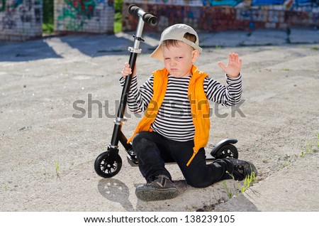 Cute camera-shy little boy sitting on the footboard of his scooter raising his hand to call a halt to the photo session while pouting with a sulky expression