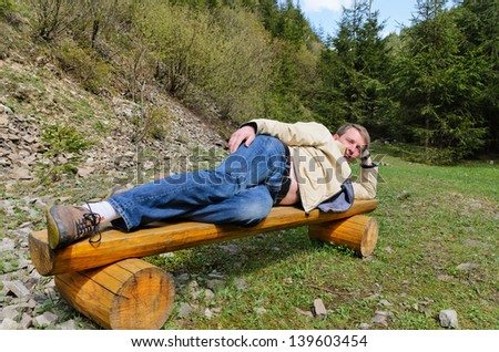 Young blond man posing for camera, stretched and laying on his side on a wooden park bench in the midst of a lush green forest.