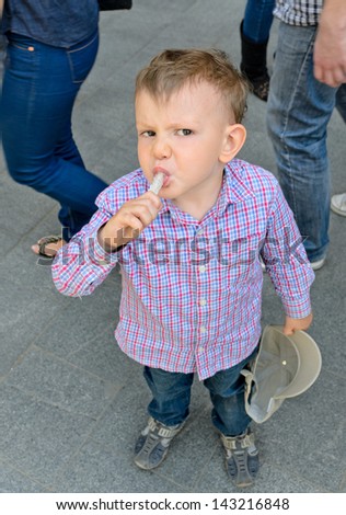 Young handsome blond little boy eating a treat in the middle of the street among people passing by
