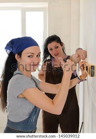 Pretty young woman giving a thumbs up of approval and success while renovating an apartment with a friend who is helping her align a spirit level on the wall