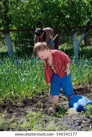 Cute little boy working in the vegetable garden with his trowel as his mother hoes weeds in the background behind him
