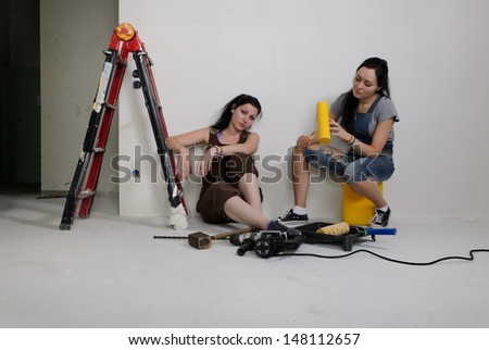 Two young women renovating a house taking a break sitting on the floor surrounded by all their tools and equipment and a stepladder