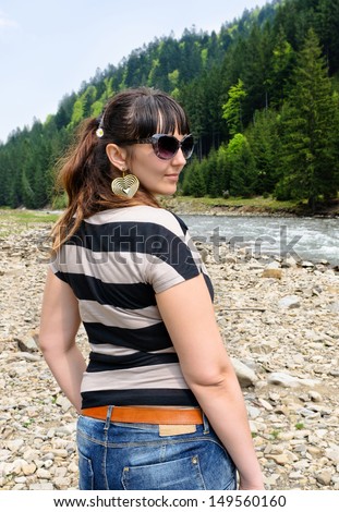 Attractive woman in a mountain valley with forested slopes and a fast flowing stream turning back to look over her shoulder