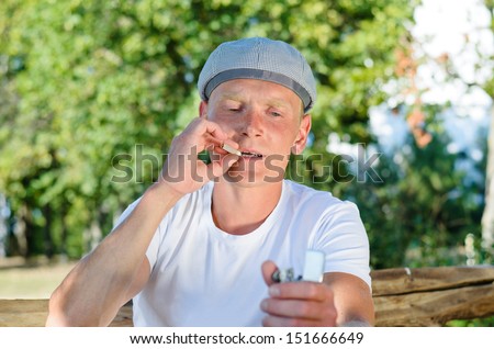 Man smoking a cigarette outdoors sitting at a wooden table puffing and inhaling as he looks at his smoking accessories in his hand for rolling the cigarette
