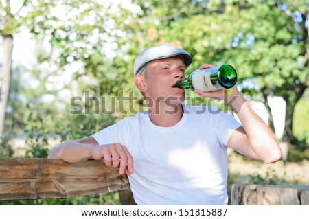 Young male alcoholic sitting alone on a wooden bench outdoors in a park drinking from a bottle of spirits