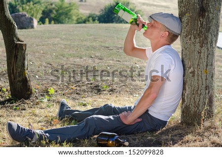 Alcoholic drinking alone in the countryside sitting on the ground with his back against a tree gulping spirits from a bottle