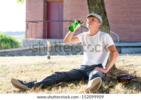 Man with an alcohol problem drinking spirits from a bottle sitting alone in a park or garden leaning against a tree in the summer sunshine