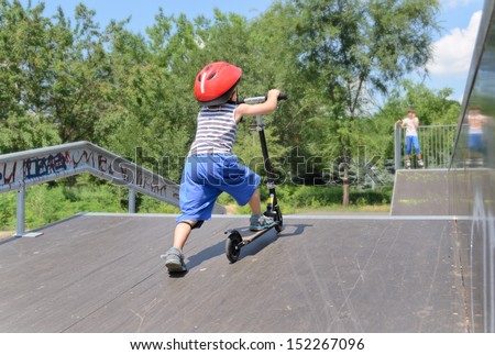 View from behind of a little boy pushing a toy scooter up a ramp at a skate park in order to use the height to gain some speed on the way down