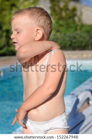 Small boy in a swimsuit at the swimming pool applying sunscreen reaching around behind his back as he rubs it into his shoulder