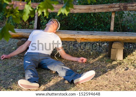 Caucasian middle-aged man lying down on the ground leaning his head on a bench feeling sick